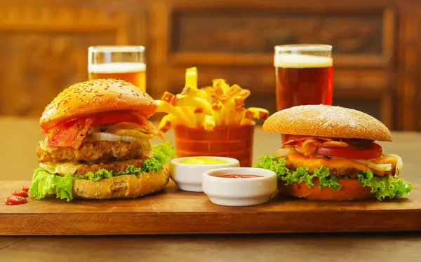 Dos deliciosas hamburguesas con ternera, cebolla, tomate, lechuga y queso con ketchup, mostaza y dos vasos de cerveza sobre tabla de madera —  Fotos de Stock