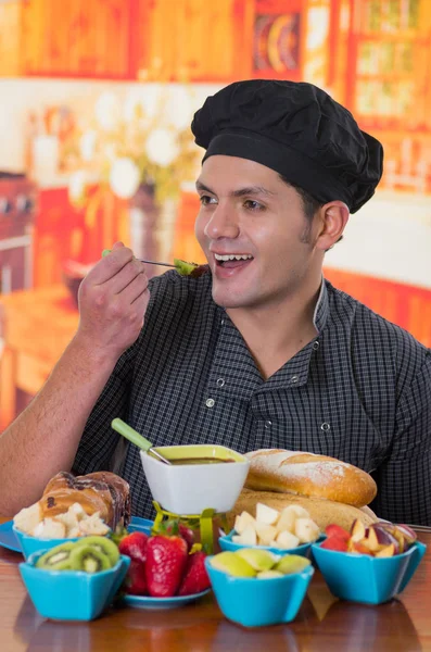 Knappe Glimlachende man eten een heerlijke kiwi bedekt met chocolade fondue binnenkant van een witte kom met diverse vruchten als, aardbei, peer, kiwi, appel en sneetjes brood in de kommen op houten tafel — Stockfoto