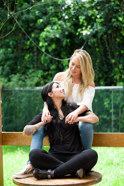 Een paar van trots lesbische in openlucht zittend op een houten tafel, blonde vrouw is een brunette vrouw, knuffelen op de achtergrond van een tuin — Stockfoto