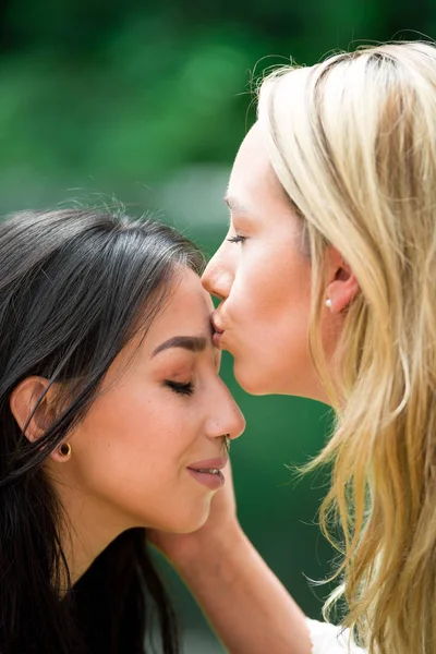 Nahaufnahme eines reizenden lesbischen Paares, blonde Frau küsst auf die Stirn eines brünetten Mädchens, vor verschwommenem Hintergrund — Stockfoto