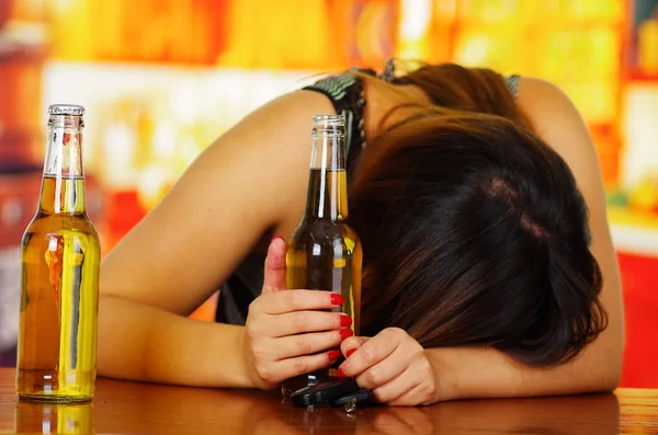 Retrato de uma mulher bêbada dormindo sobre uma mesa de madeira e segurando uma cerveja aberta com a mão, no fundo do bar — Fotografia de Stock