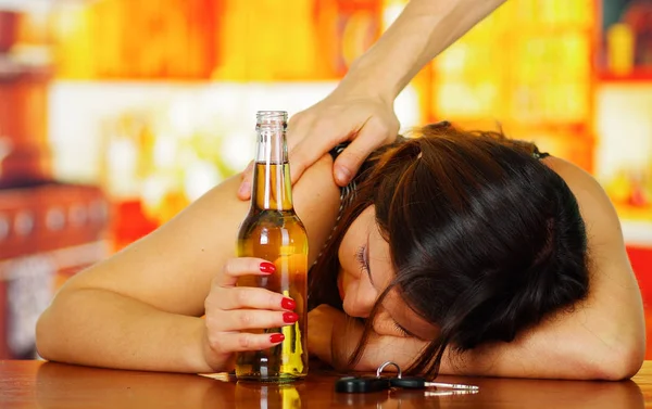 Portrait of a drunk woman sleeping over a wooden table and holding an open beer and car keys over the table, with a misterious hand in her shoulder, in bar background — Stock Photo, Image