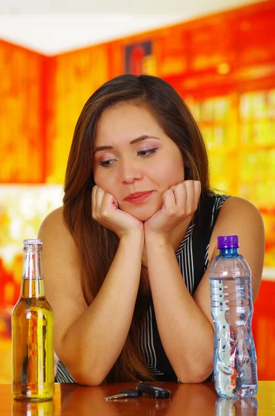 Portrait of a beautiful drunk woman posing her hands in her chin, with a bottle of beer and water and car keys over a wooden table, in bar background — Stock Photo, Image