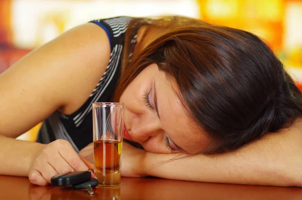 Portrait of a drunk woman sleeping over a wooden table and holding a shoot of tequila with her hand, in bar background — Stock Photo, Image