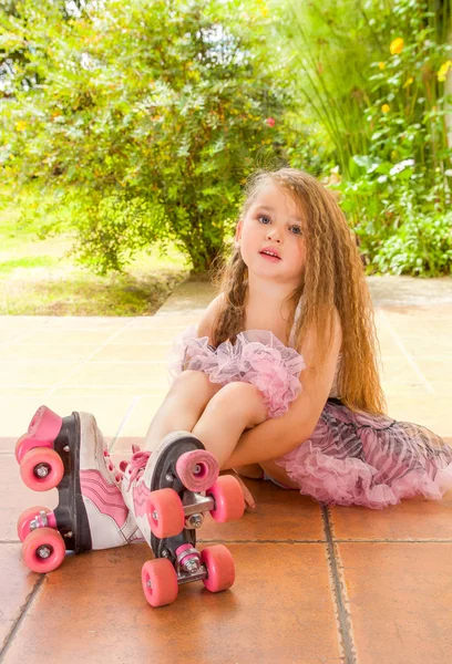 Kleine meisje preschool zittend op de grond dragen van haar rolschaatsen en overschrijding van haar benen en haar armen onder haar benen, aanbrengend een tuin achtergrond — Stockfoto