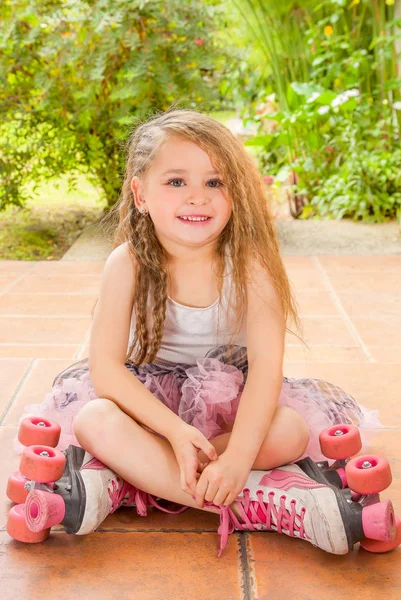 Meisje preschool zittend op de grond voor het dragen van haar rolschaatsen en het oversteken van haar benen, in een tuin achtergrond — Stockfoto