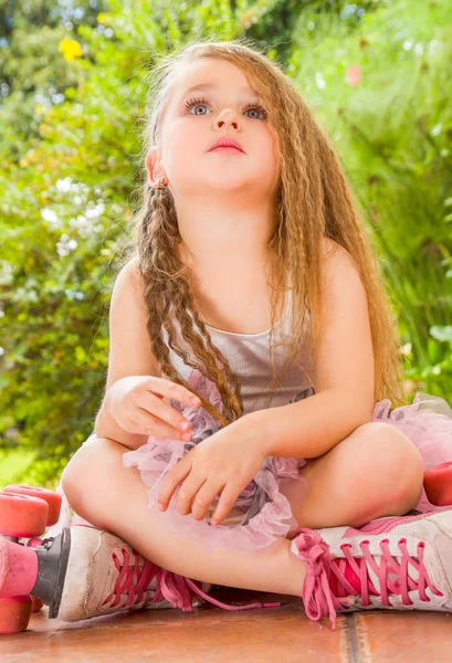 Liten flicka sitter på marken bär hennes rullskridskor och korsar benen ser himlen, i en trädgård bakgrund — Stockfoto