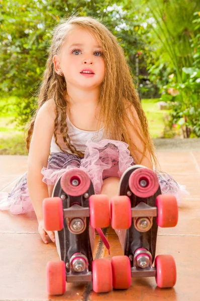 Девочка дошкольного возраста сидит на земле в роликовых коньках, на заднем плане сада — стоковое фото
