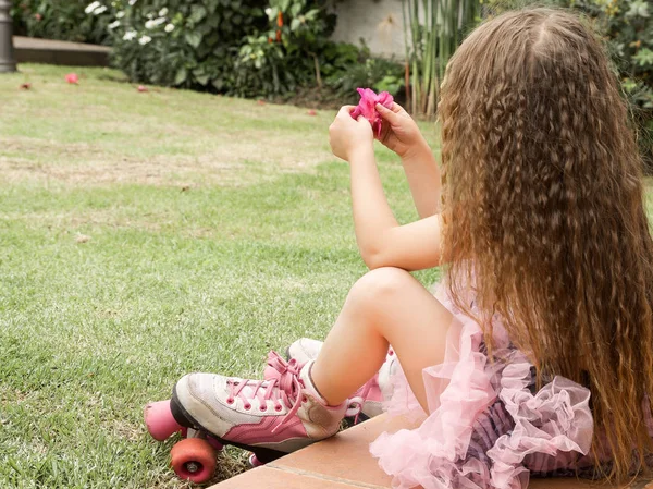 Menina pré-escolar sentada no chão usando seus patins, em um fundo de jardim, vista traseira — Fotografia de Stock