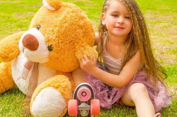 Little girl preschool sitting on backyard next to her teddy bear, wearing her roller skates and crossing her legs, in a grass background — Stock Photo, Image