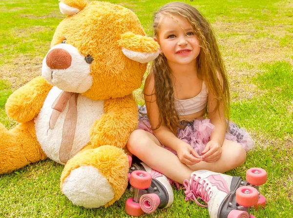 Petite fille d'âge préscolaire assise sur la cour à côté de son ours en peluche, portant ses patins à roulettes et croisant ses jambes, dans un fond d'herbe — Photo