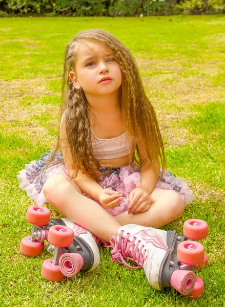 Niña preescolar sentada en el patio trasero, usando sus patines y cruzando sus piernas, en un fondo de jardín — Foto de Stock