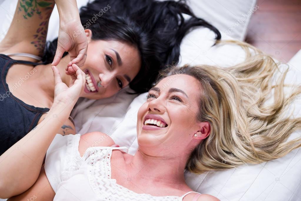 Beautiful sexy smiling lesbians lovers at morning laying in bed, doing a heart form with their hair and brunette doing with her hands a heart from, in a white background