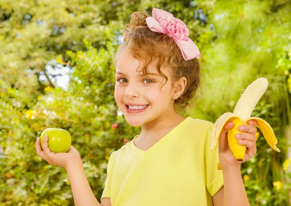 Vacker ung flicka, iklädd en gul t-shirt som håller en frisk äpple och banan, i en trädgård bakgrund — Stockfoto