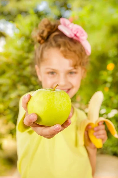 Hermosa joven con una camiseta amarilla sosteniendo un plátano saludable y señalando desde ella una manzana, en un fondo borroso jardín —  Fotos de Stock