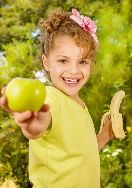 Hermosa joven con una camiseta amarilla sosteniendo un plátano saludable y señalando desde ella una manzana, en un fondo borroso jardín —  Fotos de Stock