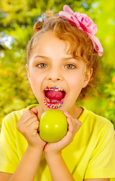 Beautiful young girl, wearing a yellow t-shirt holding a healthy apple and colorful dragees in her lips, in a blurred garden background — Stock Photo, Image