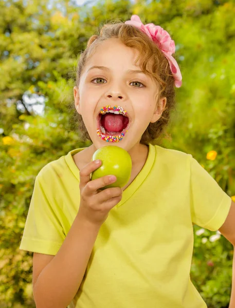 Hermosa joven, vistiendo una camiseta amarilla y comiendo una manzana sana y coloridos dragees en sus labios, en un fondo borroso jardín —  Fotos de Stock