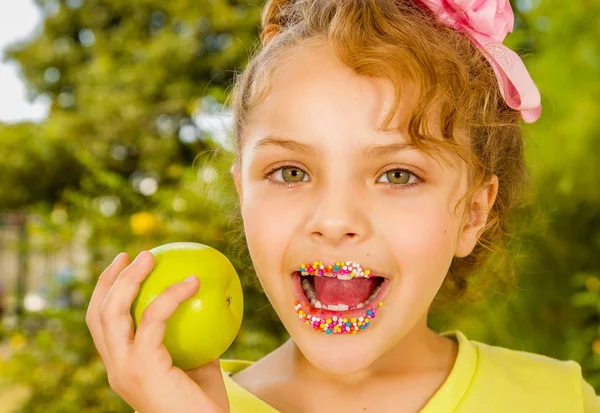 Acercamiento de una hermosa joven, con una camiseta amarilla sosteniendo una manzana sana y coloridos dragees en sus labios, en un fondo borroso jardín —  Fotos de Stock