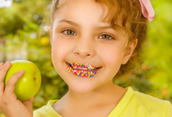 Close up of a beautiful young girl, wearing a yellow t-shirt holding a healthy apple and colorful dragees in her lips, in a blurred garden background — Stock Photo, Image