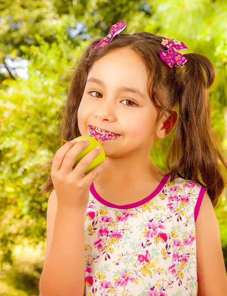 Beautiful young girl, eating a healthy apple and colorful dragees in her lips, in a blurred garden background — Stock Photo, Image