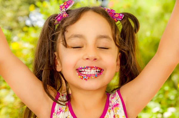 Close up of a beautiful young girl, with colorful dragees in her lips with both arms up, in a blurred garden background — Stock Photo, Image
