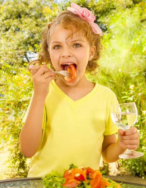 Beautiful young girl, wearing a yellow t-shirt eating a tomato from her salad and holding glass of water, in a garden background — Stock Photo, Image