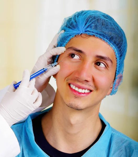 Gros plan d'un beau jeune homme souriant, recevant des injections de traitement cosmétique du visage, la main des médecins avec une seringue tenant un gant et portant un chapeau médical — Photo