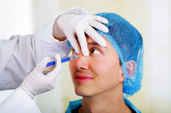 Close-up de um belo jovem sorridente, recebendo injeções de tratamento cosmético facial em seu nariz, médicos mão com luva segurando seringa e vestindo um chapéu médico usando um chapéu de cirurgia — Fotografia de Stock