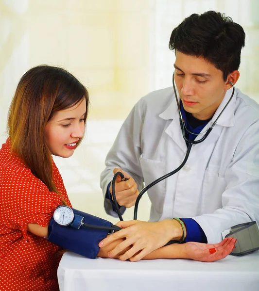 Närbild av en stilig ung läkare med stetoskopet runt halsen tar pulsen till en ung kvinna med en tensiometer, i en läkare samråd rum bakgrund — Stockfoto