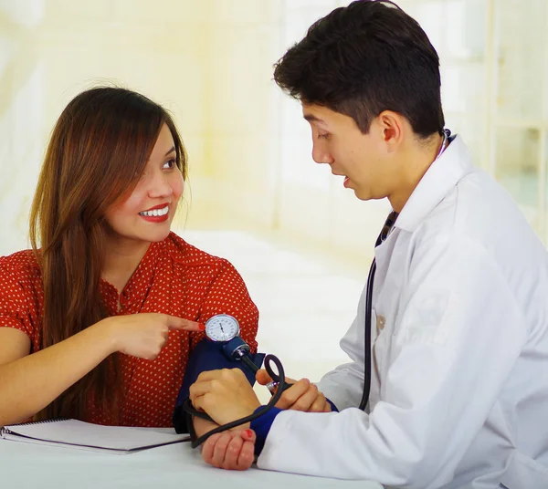 Bonito surpreendido jovem médico com estetoscópio em torno de seu pescoço levando o pulso para uma jovem mulher, em um médico consultório fundo — Fotografia de Stock