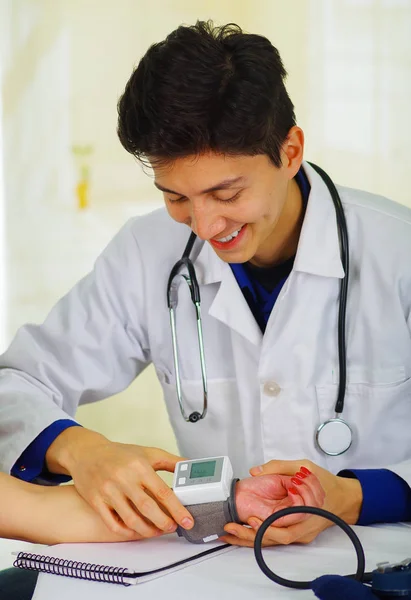 Bonito sorridente jovem médico com estetoscópio em torno de seu pescoço levando o pulso para uma jovem mulher, em um médico consultório fundo — Fotografia de Stock