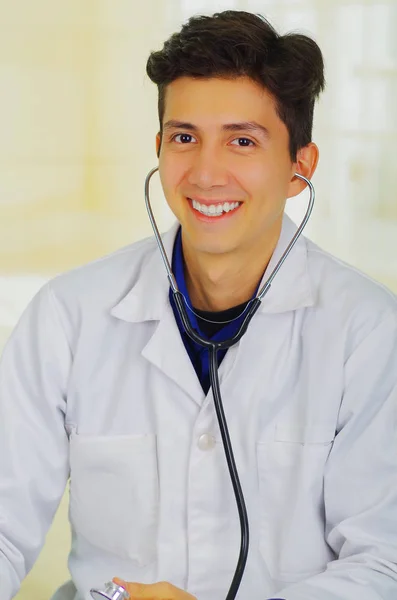 Красивый улыбающийся молодой доктор со стетоскопом вокруг шеи, выступающий перед камерой, в кабинете врача — стоковое фото