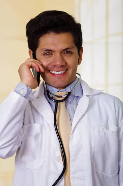 英俊的笑脸医生用听诊器抱着他的脖子上，在办公室背景中使用他的 celphone — 图库照片