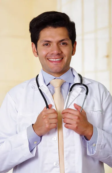 Confiado guapo sonriente médico posando y mirando a la cámara con un estetoscopio alrededor de su cuello, en el fondo de la oficina — Foto de Stock