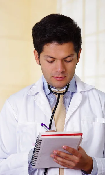 Médico guapo con un estetoscopio sosteniendo de su cuello, escribiendo en sus notas de cuaderno en el fondo de la oficina — Foto de Stock