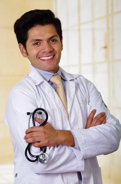 Красивый улыбающийся доктор позирует и смотрит в камеру со стетоскопом в руках со скрещенными руками, на офисном фоне — стоковое фото