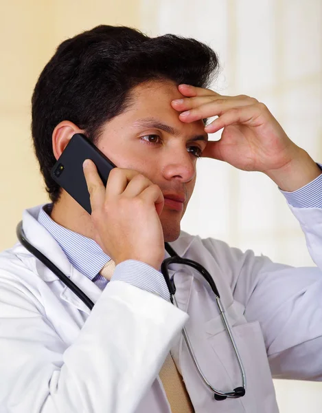 Уверенный в себе красивый врач со стетоскопом на шее, с помощью телефона одной рукой и другой рукой он прикасается к голове, на офисном фоне — стоковое фото