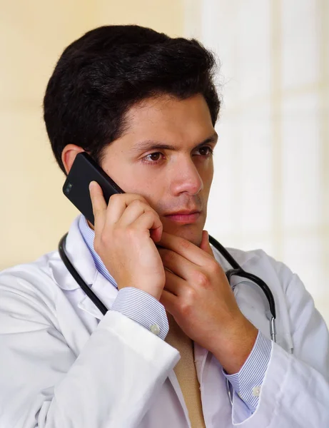Selbstbewusster, gutaussehender Arzt mit Stethoskop um den Hals, der ein Handy mit einer Hand benutzt, während er sein Kinn mit der anderen Hand berührt, im Hintergrund im Büro — Stockfoto