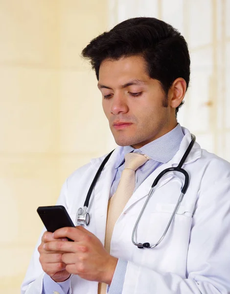 Médico bonito confiante com um estetoscópio ao redor do pescoço, usando um celular com ambas as mãos, no fundo do escritório — Fotografia de Stock