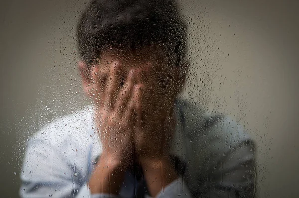 Worry depresed young man, hiding from camera using his hands, behind a blurred window with drops, gray background