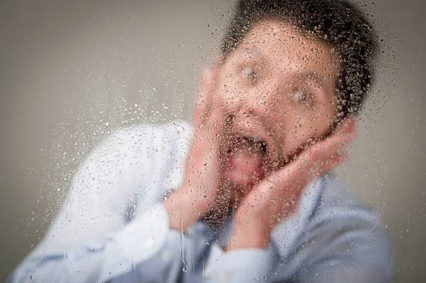 Junger Mann schreit mit beiden Händen im Gesicht hinter einem verschwommenen Fenster mit Tropfen, grauer Hintergrund — Stockfoto