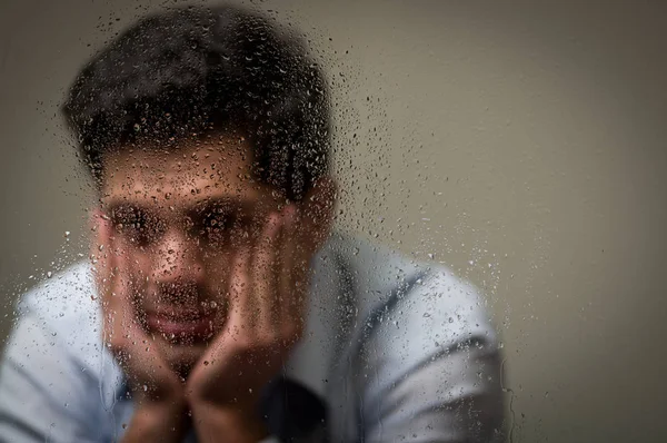 Preocupe-se jovem deprimido com as duas mãos no rosto, atrás de uma janela embaçada com gotas, fundo cinza — Fotografia de Stock
