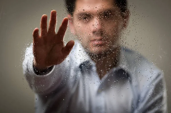 Young business man, behind a blurred window with drops, pointing in from of him an open hand, gray background — Stock Photo, Image