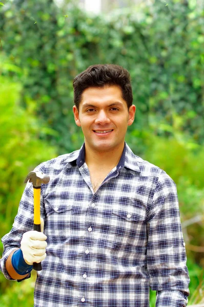 Atractivo hombre con ropa casual sosteniendo un martillo en un fondo del bosque — Foto de Stock