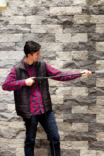 Όμορφος νεαρός άνδρας φορώντας τετράγωνο μοτίβο κόκκινο κρατώντας υψηλή πίεση νερού όπλο, δείχνει προς το γκρίζο τοίχο — Φωτογραφία Αρχείου
