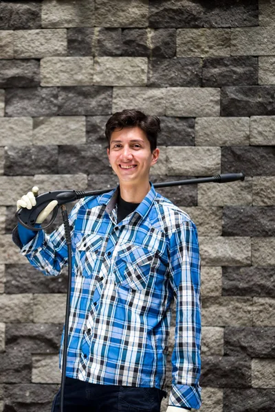 Handsome young man wearing square pattern blue holding on his shoulder a high pressure water gun, on a garden background — Stock Photo, Image