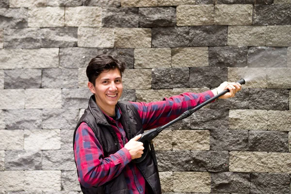 Hombre joven guapo con patrón cuadrado rojo sosteniendo pistola de agua de alta presión, apuntando hacia la pared de ladrillo gris — Foto de Stock