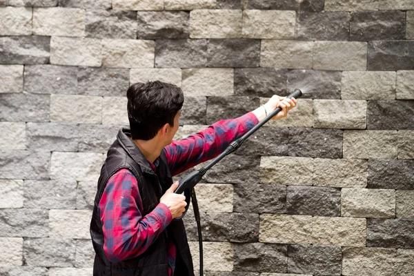 Bonito jovem vestindo padrão quadrado vermelho segurando arma de água de alta pressão, apontando para a parede de tijolo cinza — Fotografia de Stock
