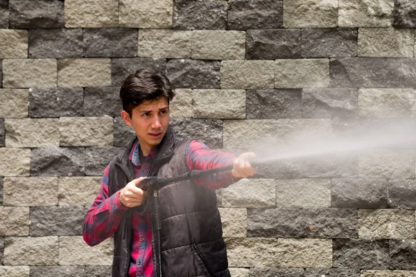 Hombre joven guapo con patrón cuadrado rojo sosteniendo pistola de agua de alta presión, sobre un fondo de pared de ladrillo gris — Foto de Stock